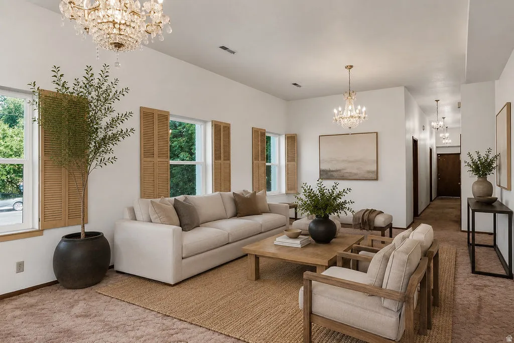 Spacious living area featuring dual crystal chandeliers, multiple windows with natural wood shutters, a neutral color palette, and high ceilings