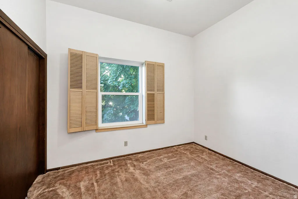 Room featuring a window with louvered shutters, white walls, and brown carpeting