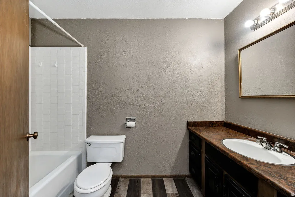 Bathroom featuring a white tub and shower surround with tile, a vanity with a white oval sink, a brown patterned countertop, dark wood-finish cabinetry, and wood-finish flooring