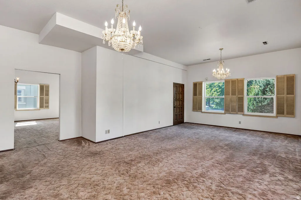 Spacious room featuring two ornate chandeliers, ample natural light from multiple windows, and a dark wood-finish entry door