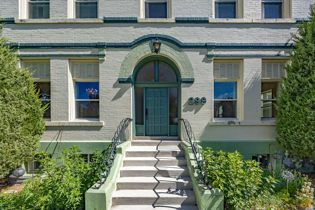 Brick facade featuring a prominent arched entryway with a teal door, concrete steps with decorative wrought iron railings, and mature landscaping