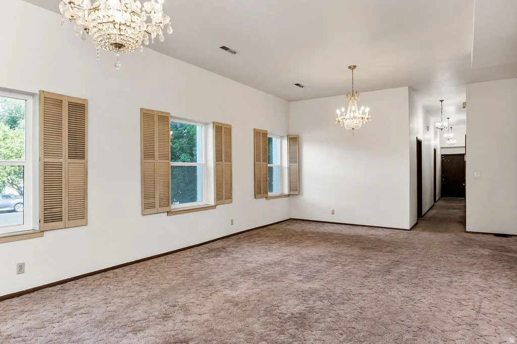 Spacious interior room featuring two crystal chandeliers, three windows with natural wood louvered shutters, and textured wall-to-wall carpeting