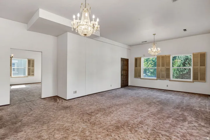 Spacious room featuring two ornate chandeliers, ample natural light from multiple windows, and a dark wood-finish entry door