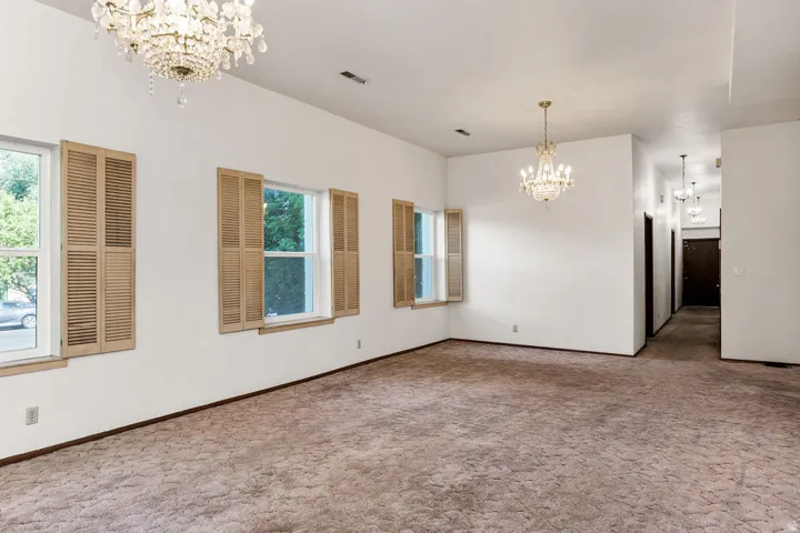 Spacious interior room featuring two crystal chandeliers, three windows with natural wood louvered shutters, and textured wall-to-wall carpeting
