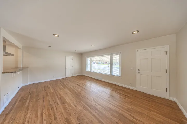 Spacious interior featuring wood flooring, recessed lighting, a large picture window with blinds, a six-panel entry door, and a built-in counter with an overhead stainless steel range hood