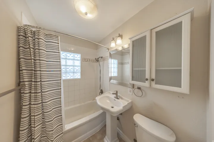 Bathroom featuring a pedestal sink, white tile shower surround with glass block window, and dual wall-mounted storage cabinets with reeded glass inserts
