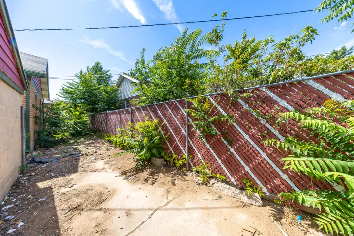 Exterior side yard space with a combination of concrete and dirt ground cover, featuring a red and white privacy fence and mature green foliage