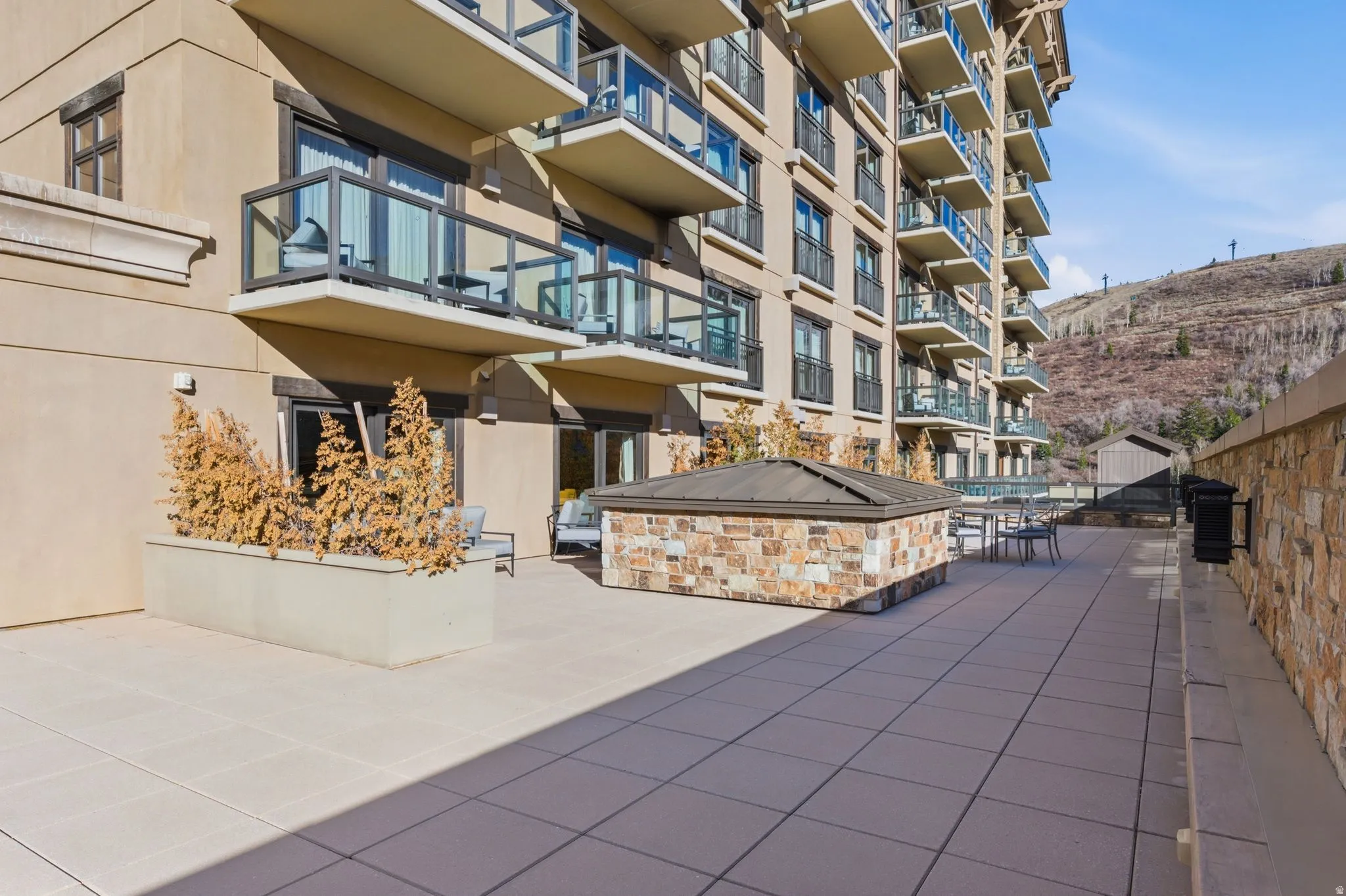 Expansive outdoor terrace featuring large format pavers, a stone-clad built-in structure with a metal roof, and a stone retaining wall