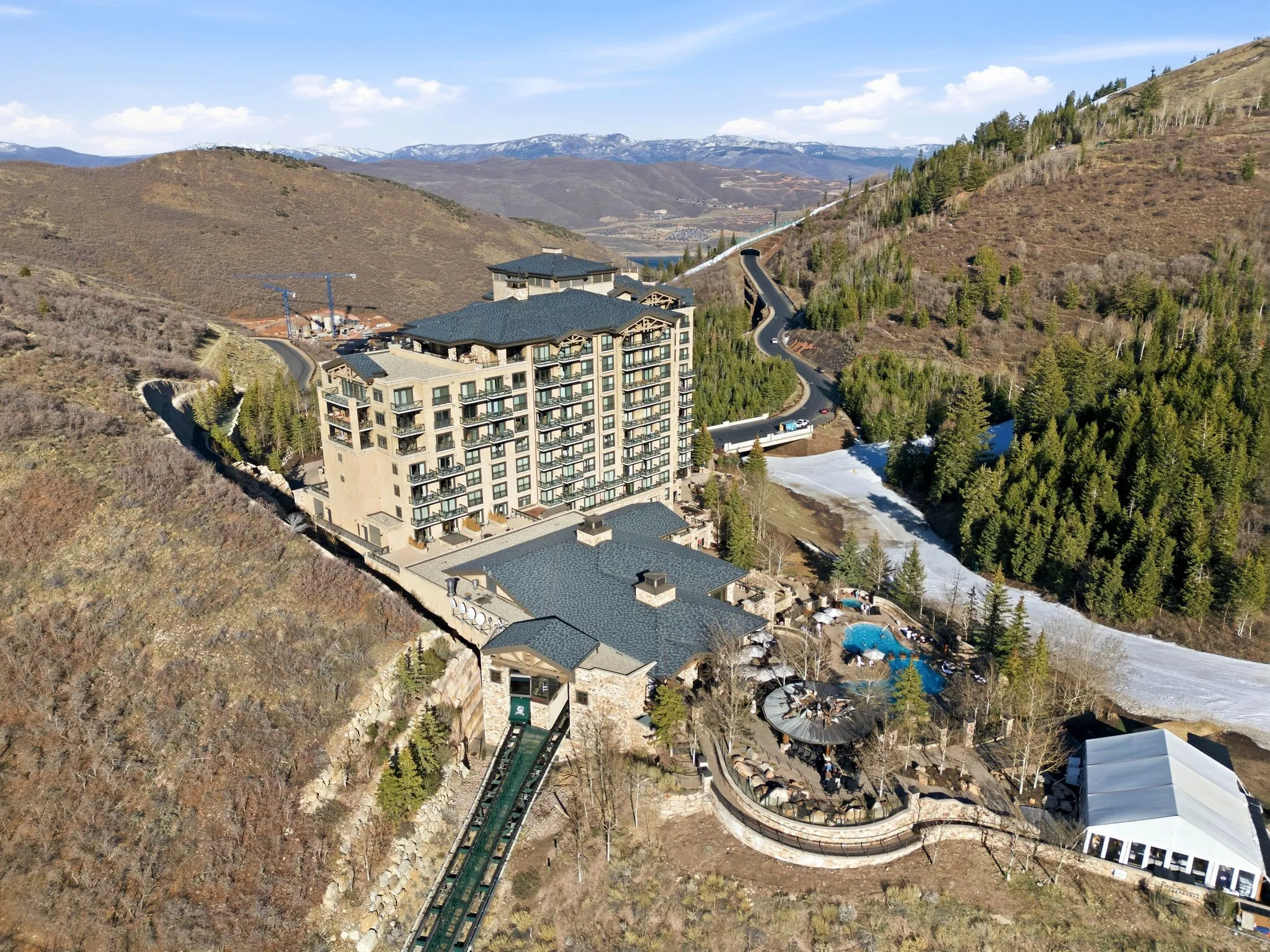 Multi-story resort-style building featuring numerous private balconies, a terraced outdoor pool area with integrated hot tubs, a private funicular lift, and surrounding mountainous terrain with snow-covered sections