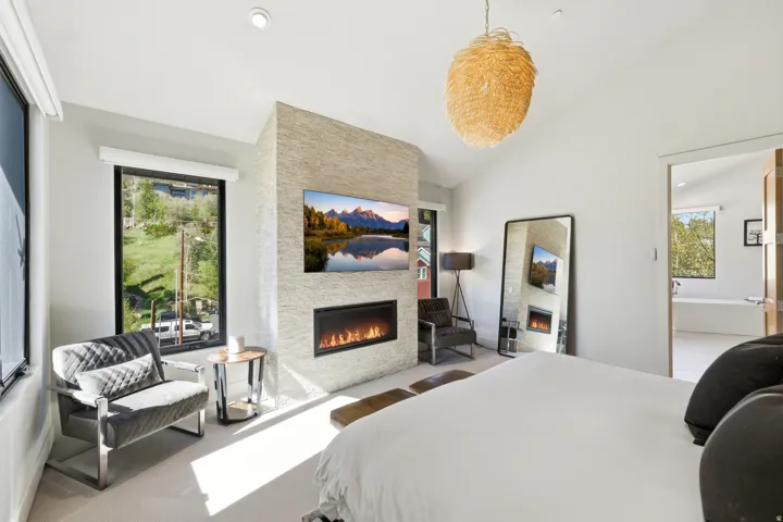Vaulted ceiling bedroom featuring a stone-clad fireplace wall with an integrated linear fireplace, a wall-mounted television, and a large window with roller shades