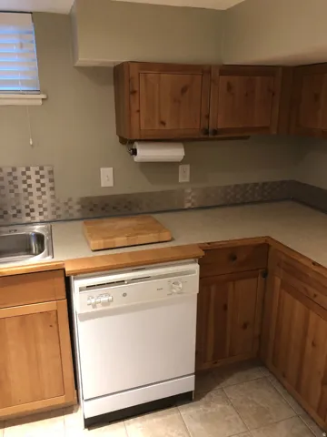 Kitchen area featuring wood cabinetry, a stainless steel sink, a white dishwasher, light-toned countertops, and a tiled backsplash