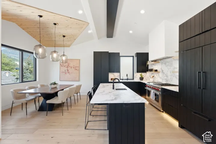 Open-concept kitchen and dining area featuring light wood-finish flooring, a vaulted ceiling with exposed wood paneling, black cabinetry, white countertops, and a full-height marble-patterned backsplash