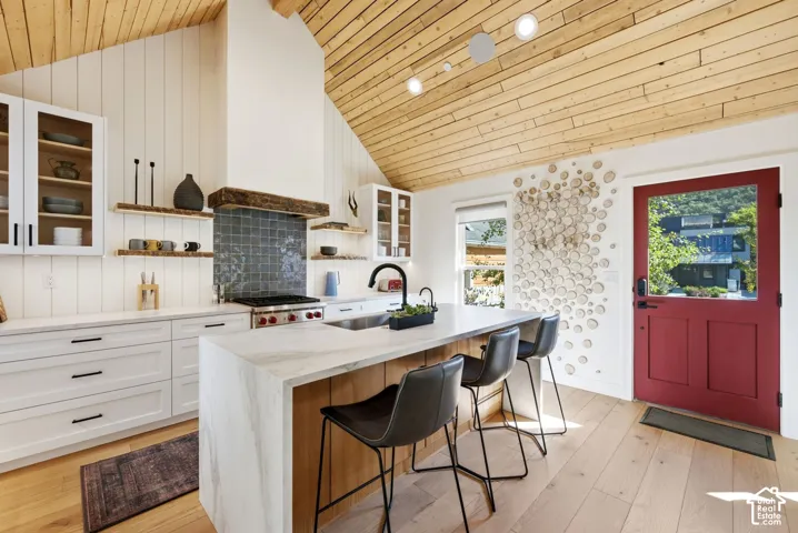 Modern kitchen featuring a vaulted wood-plank ceiling, waterfall island with stone-finish countertop, white flat-panel cabinetry, and light wood-finish flooring
