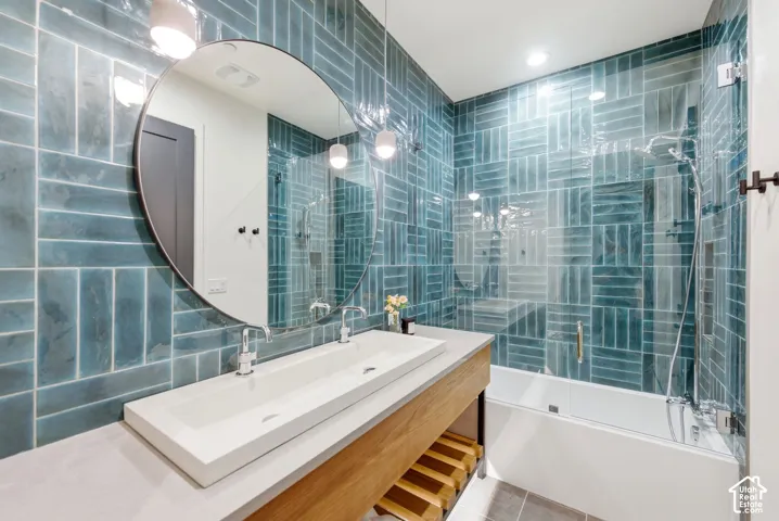 Bathroom featuring full-height teal subway tile, a vessel sink with dual faucets, a circular mirror, and a built-in shower/tub combination with a glass partition