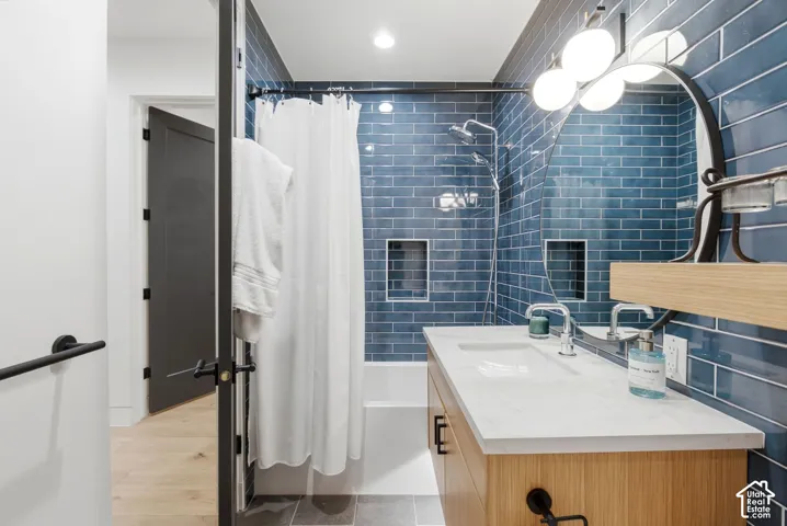 Bathroom featuring a wood-finish vanity with a light-toned countertop, a shower-over-tub with blue subway tile surround, two tiled shower niches, and a circular wall mirror with an integrated light fixture