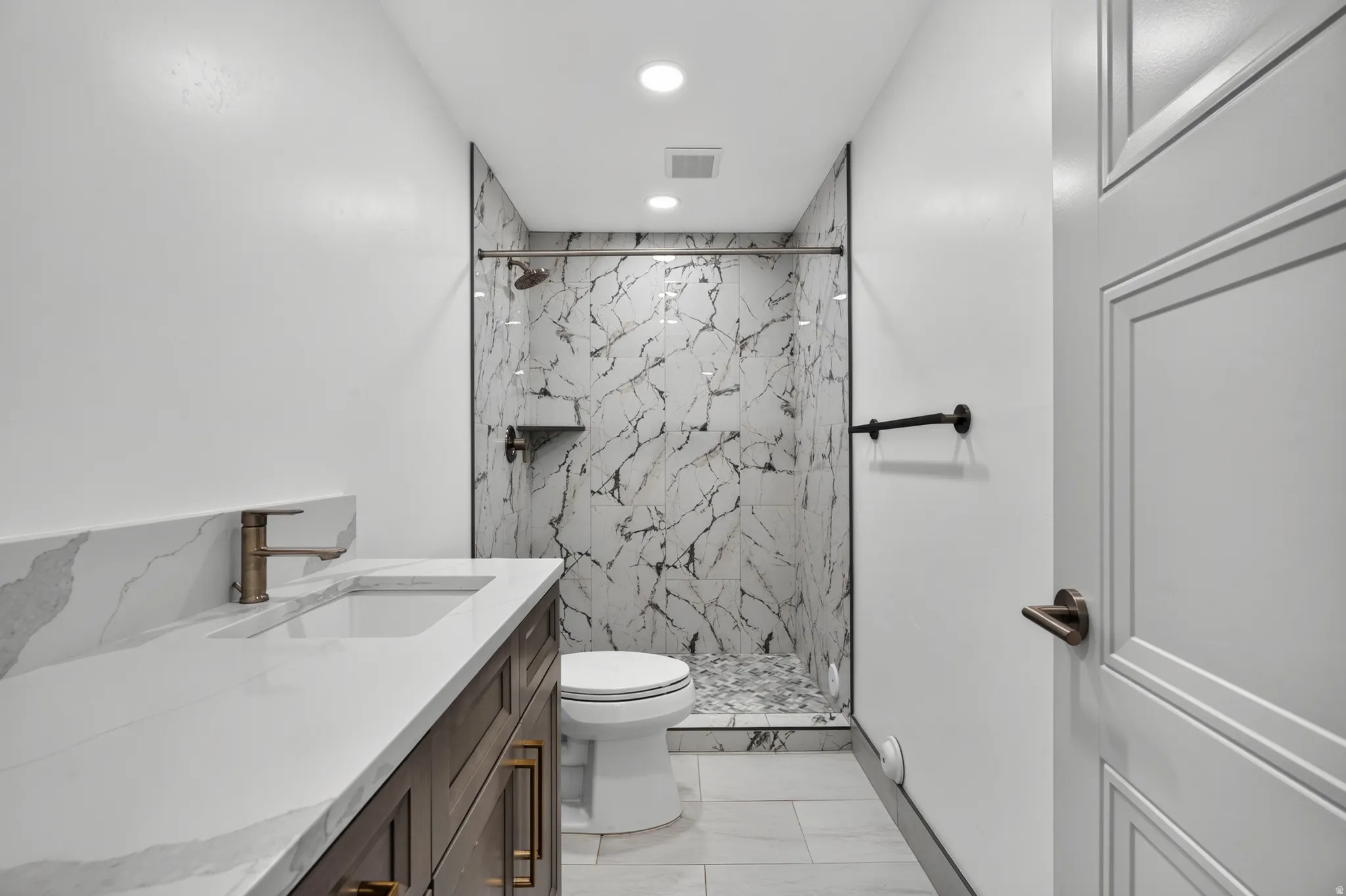 Contemporary bathroom featuring a dark wood-finish vanity with a white countertop and integrated sink, bronze-finish faucet, and a walk-in shower with black-veined white tile walls