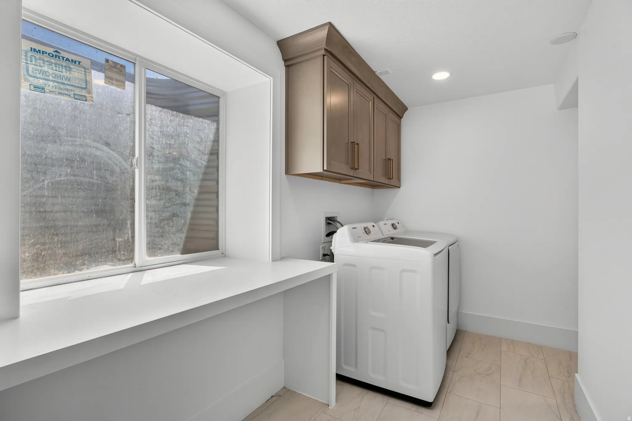 Laundry area with a window, white walls, light-toned tile flooring, and built-in cabinetry with gold-toned hardware