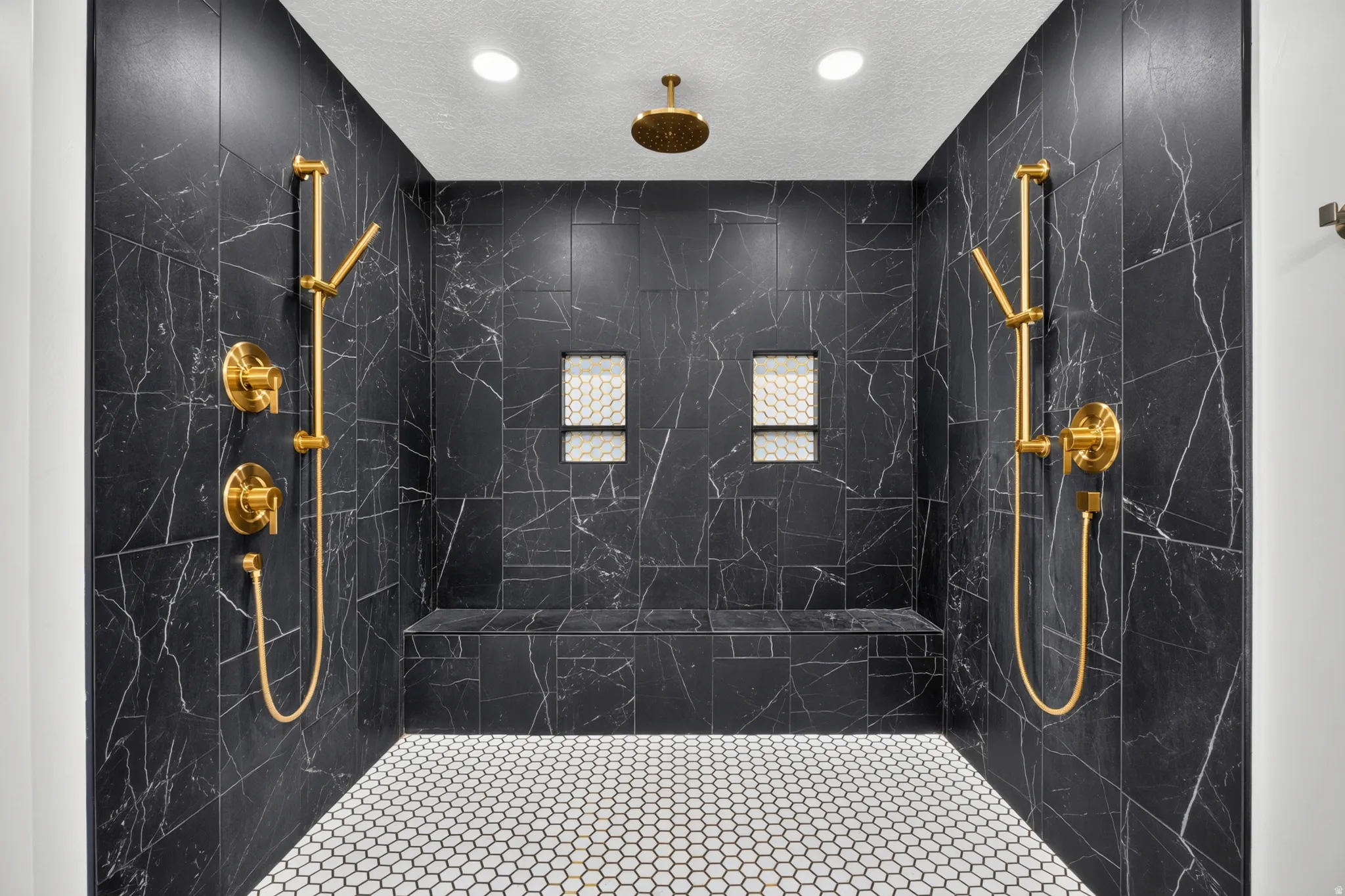 Walk-in shower featuring black tile walls with white veining, dual handheld shower heads, a rain shower head, and a built-in bench
