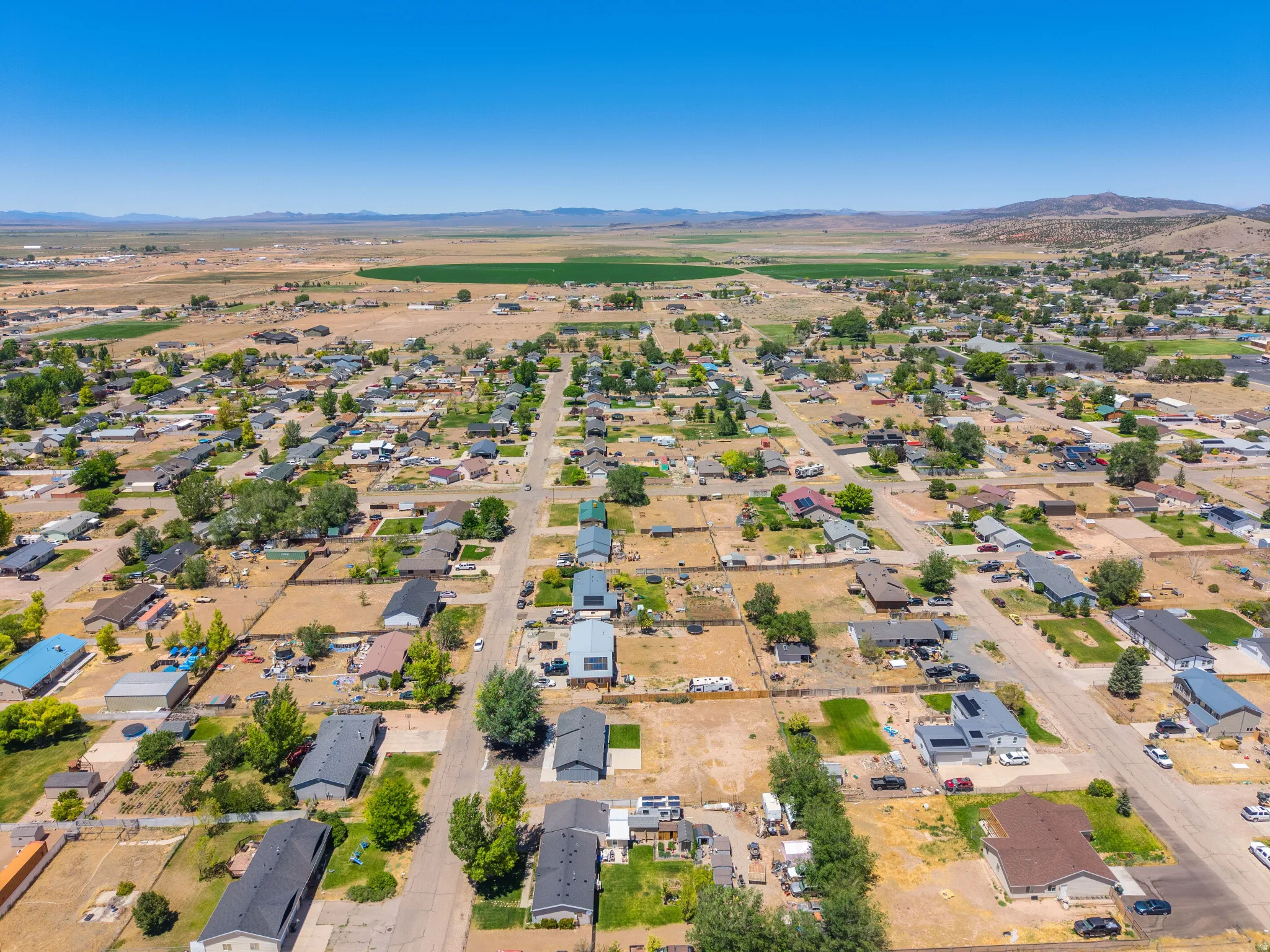 Aerial perspective showcasing a residential neighborhood with a mix of single-story homes and established trees, set against a backdrop of distant mountains and agricultural fields