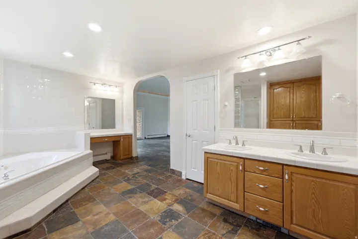 Spacious bathroom featuring a dual vanity with oak cabinetry and white countertops, a large jetted tub with tile surround, and an arched doorway leading to an additional space
