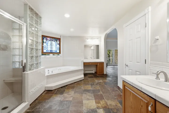 Spacious bathroom featuring natural stone flooring, a glass-enclosed shower with built-in bench, a jetted soaking tub, and dual vanity areas with solid surface countertops