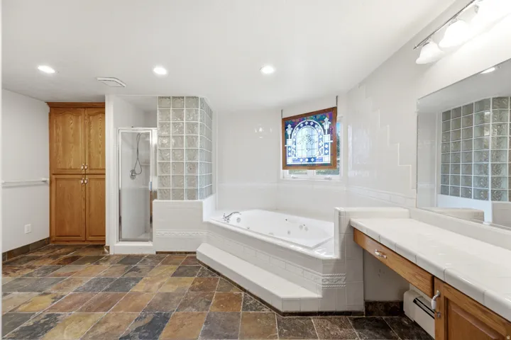 Spacious bathroom featuring a multi-toned slate tile floor, a built-in jetted tub, a glass-block shower enclosure, a stained-glass window, and a wood-finish vanity with a tile countertop