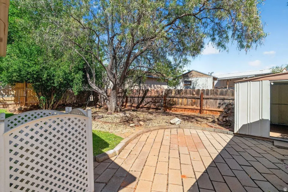Expansive outdoor patio area featuring brick paver flooring, a white lattice fence, a large mature tree, a storage shed, and a privacy fence