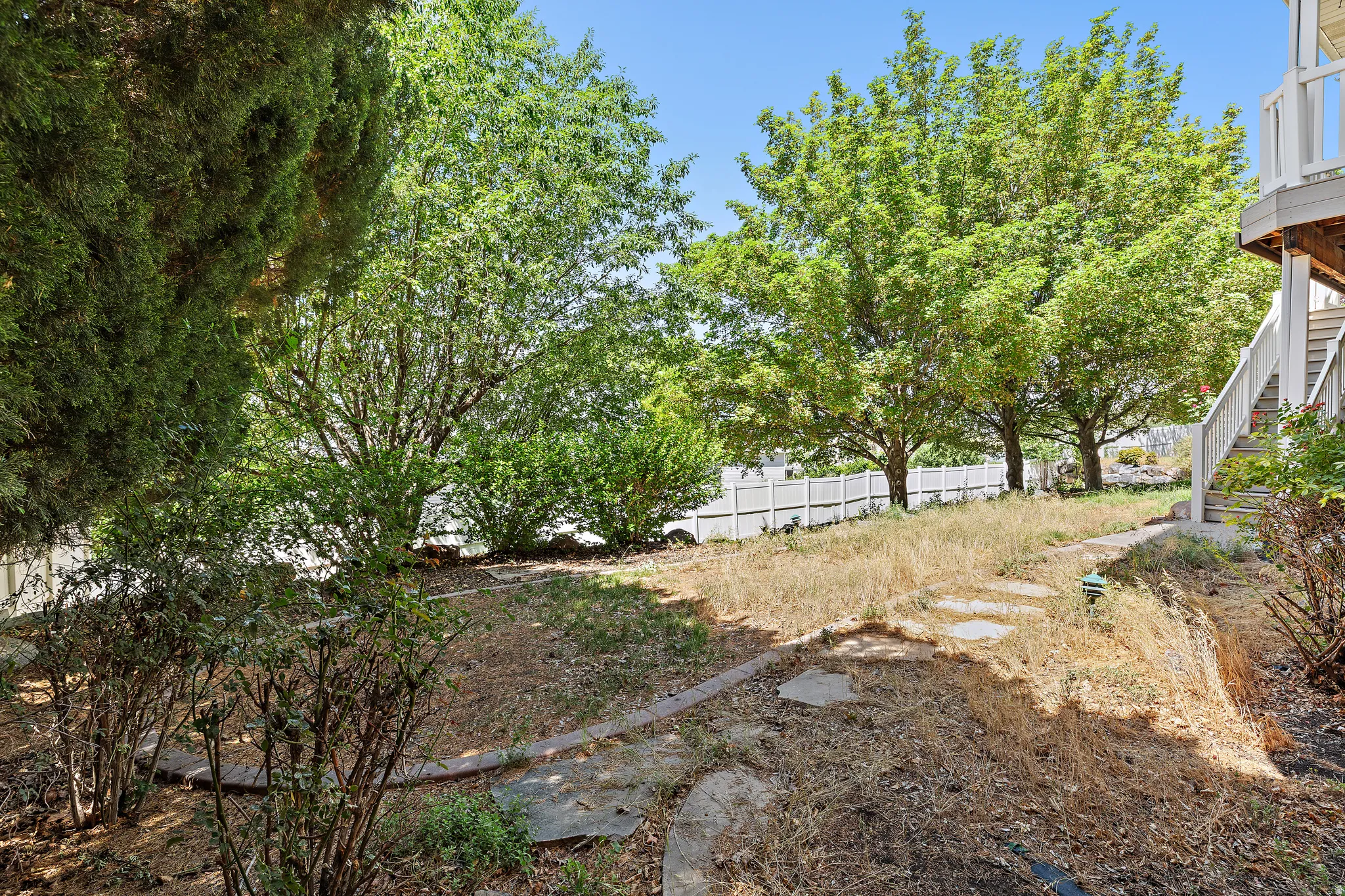 Tiered backyard featuring mature trees, established shrubs, a white privacy fence, and concrete stepping stones
