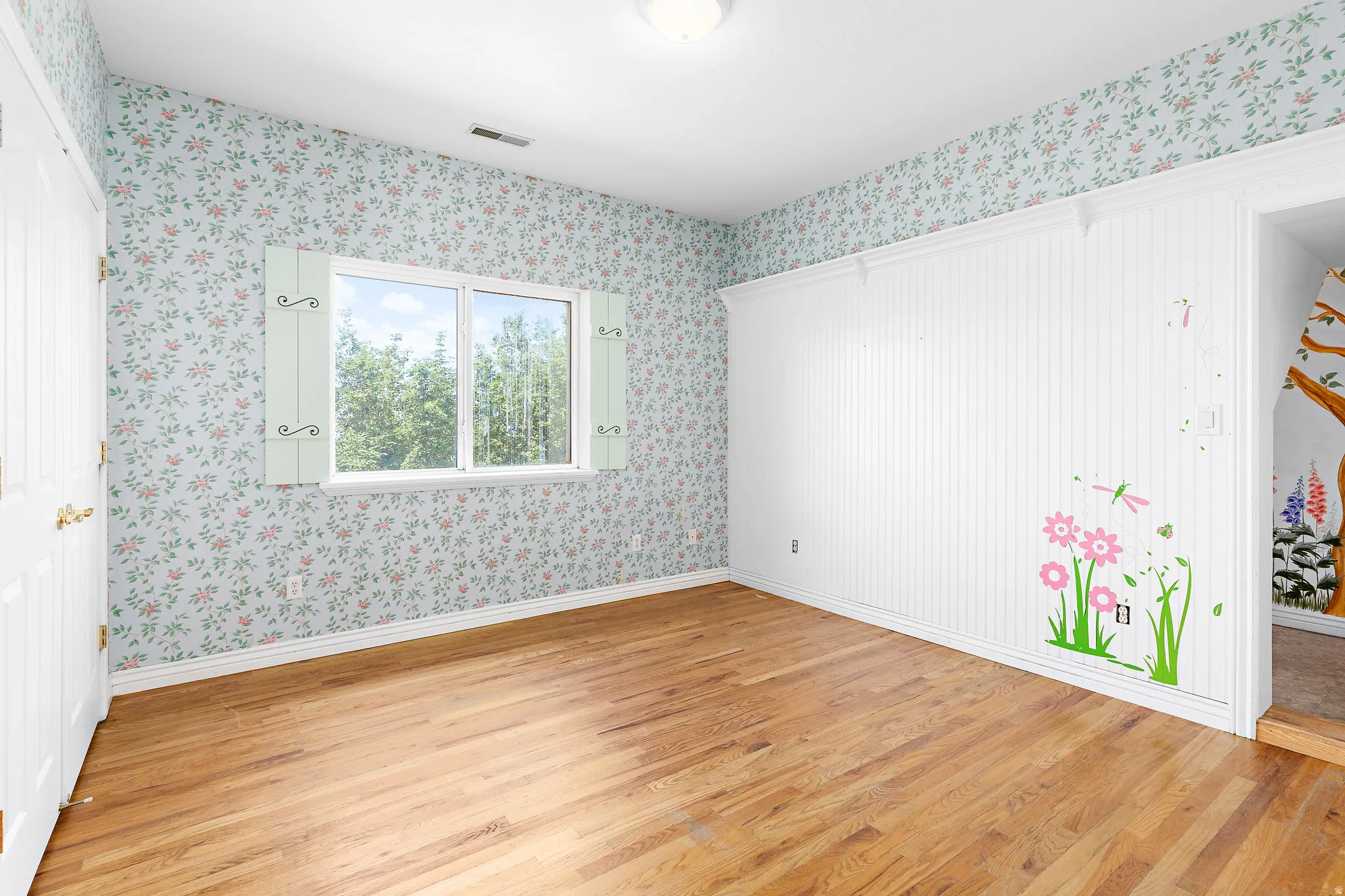 Room with hardwood flooring and patterned wallpaper, featuring a window with decorative shutters