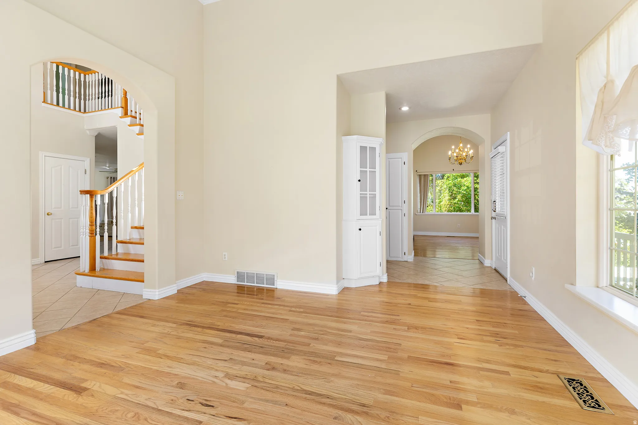 Bright interior featuring hardwood flooring, arched doorways, a built-in corner cabinet, a window with exterior visibility, and a visible staircase with wood newel posts and handrails