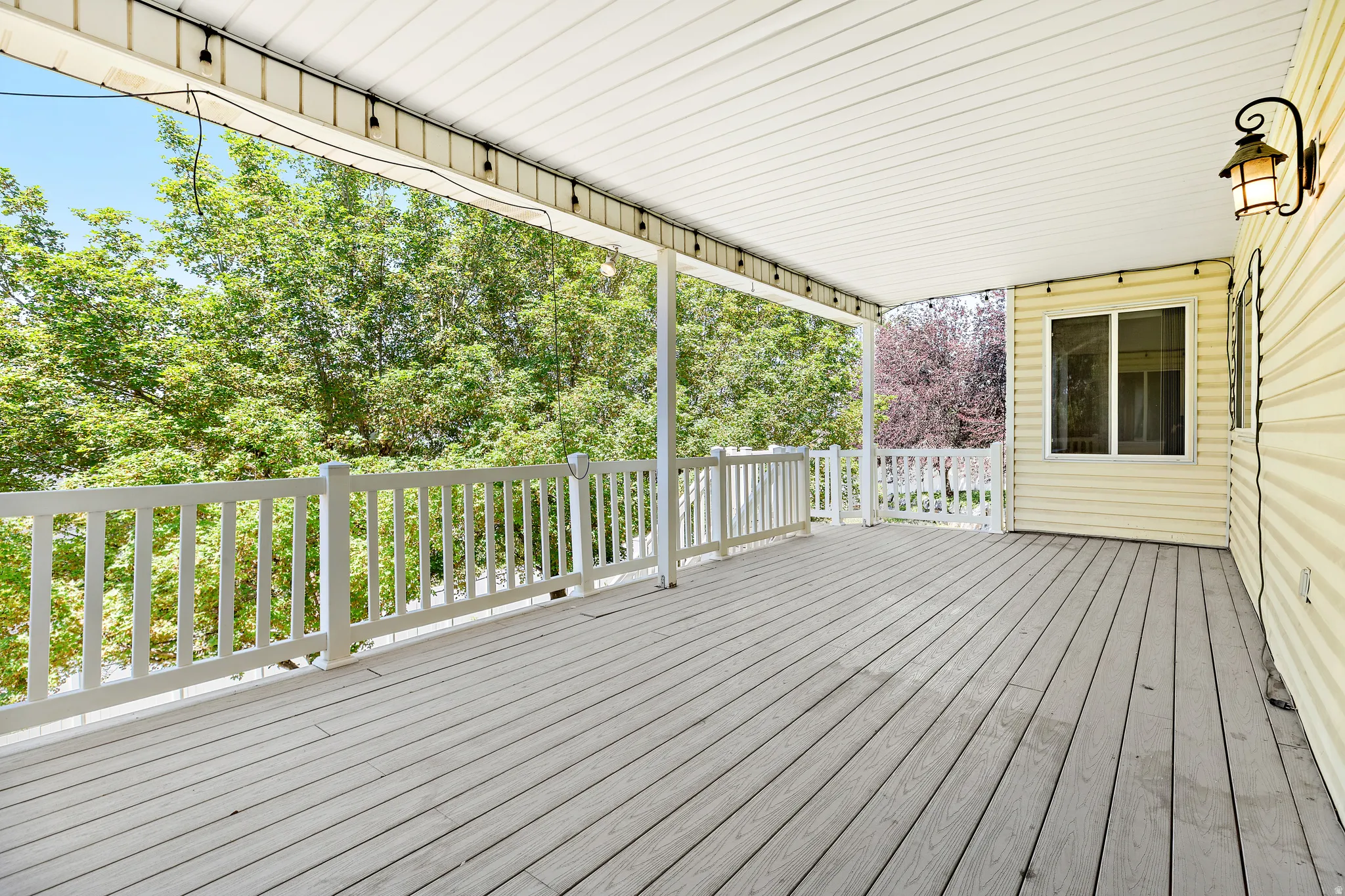 Spacious covered deck featuring wood-finish flooring, white railing, a fixed window, and an exterior wall light fixture