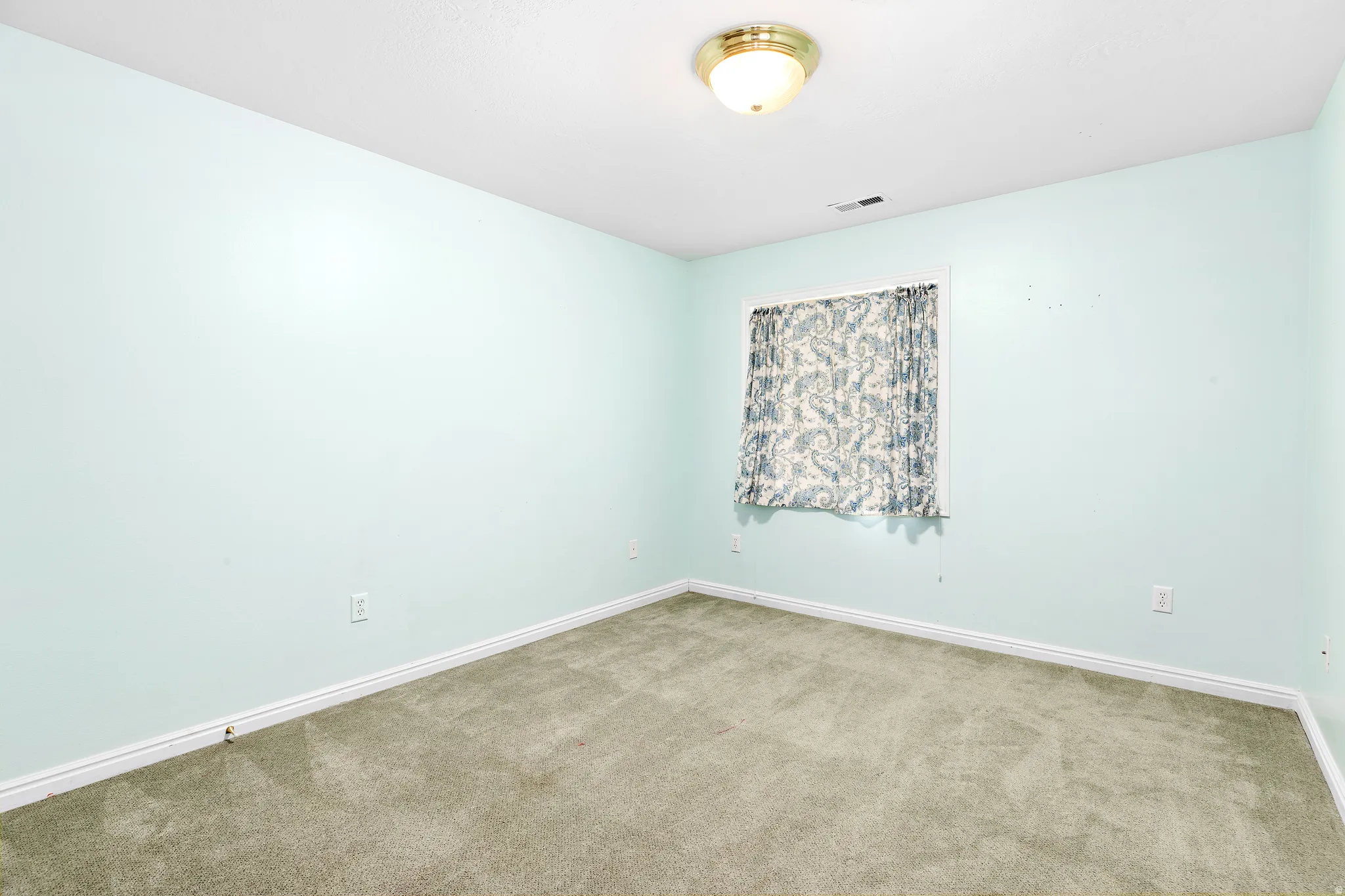 Carpeted room featuring a single window with patterned drapes, light blue walls, and white baseboards