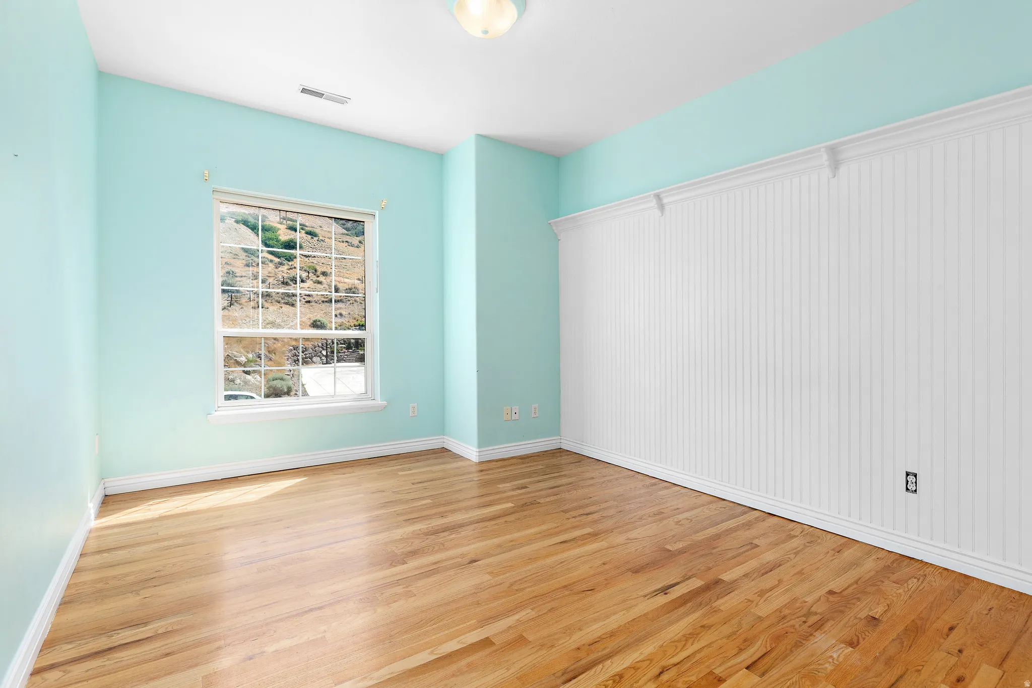 Room featuring wood-finish flooring, a window with exterior scenery, a half-wall of white beadboard paneling with decorative trim, and light blue painted walls