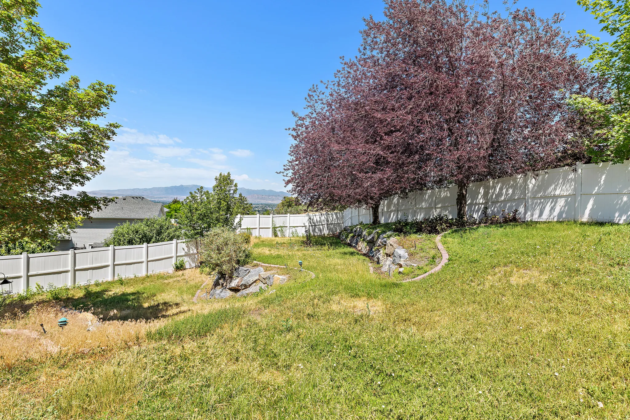 Expansive backyard featuring a lush lawn, mature trees, white privacy fencing, and a terraced landscape with natural rock elements