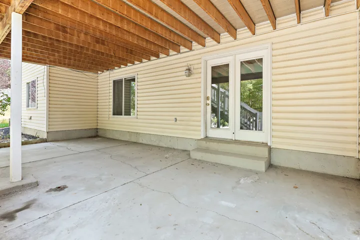 Covered concrete patio space featuring exposed wood joists overhead, light-colored horizontal siding, and a double-pane glass door entrance with a two-step concrete landing