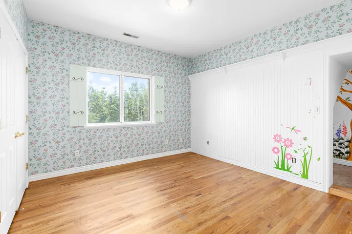 Room with hardwood flooring and patterned wallpaper, featuring a window with decorative shutters