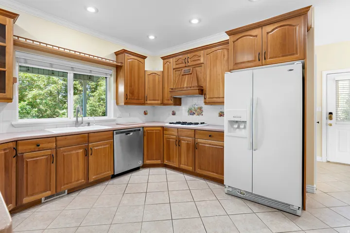 Kitchen featuring wood cabinetry, white tile backsplash, corner sink with a stainless steel faucet, stainless steel dishwasher, and tile flooring
