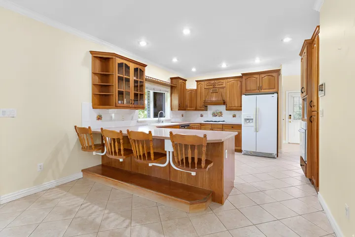 Spacious kitchen featuring warm wood cabinetry, light-toned tile flooring, recessed lighting, a built-in breakfast bar, and a double-door refrigerator