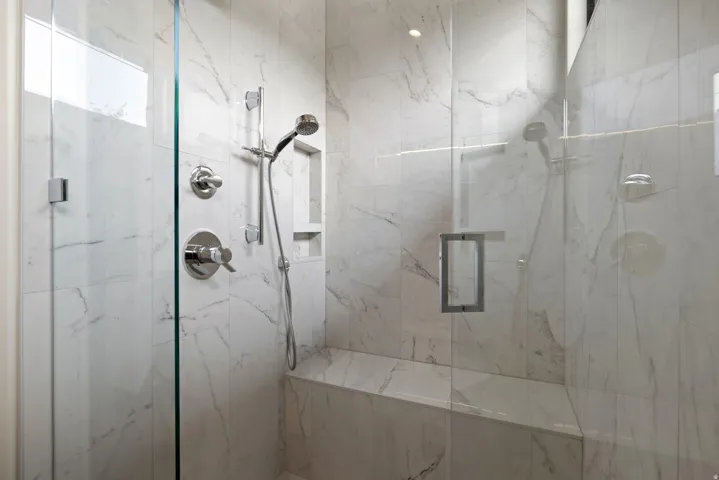 Walk-in shower featuring clear glass enclosure, marble-look tile walls, chrome fixtures, built-in bench, and recessed storage niche