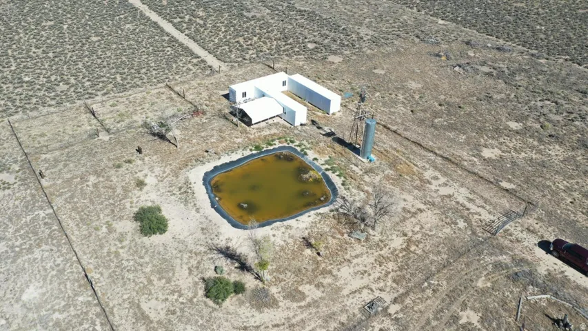 Aerial view of a secluded property featuring a white contemporary structure, an artificial pond, and a tall metal structure, all surrounded by arid terrain