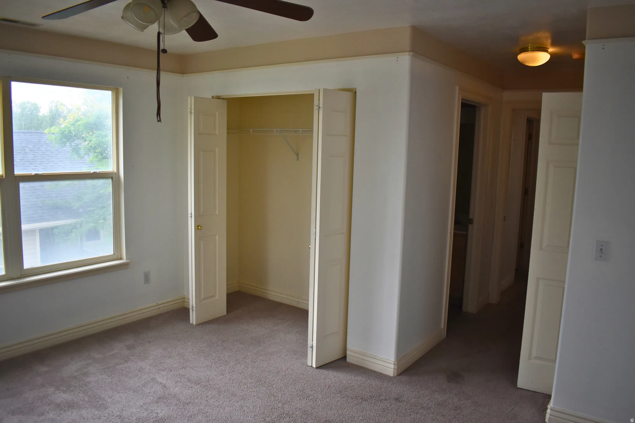 Carpeted bedroom featuring a ceiling fan with light fixture, large window, and bi-fold closet doors with wire shelving