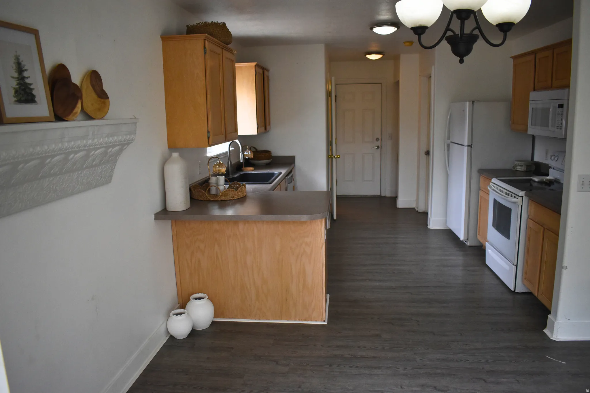 Kitchen area featuring wood-finish flooring, light wood cabinetry, white appliances, dark countertops, and a peninsula with seating space