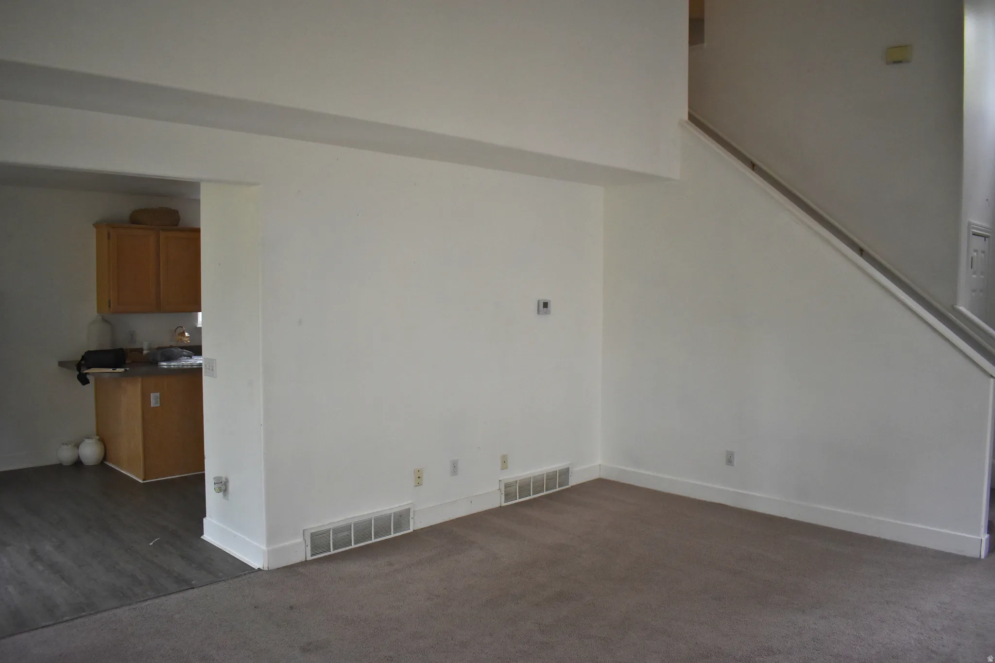 Carpeted living area with light-colored walls, a staircase with a handrail, and a pass-through opening to a kitchen space featuring wood-finish cabinetry, a countertop, and wood-finish flooring