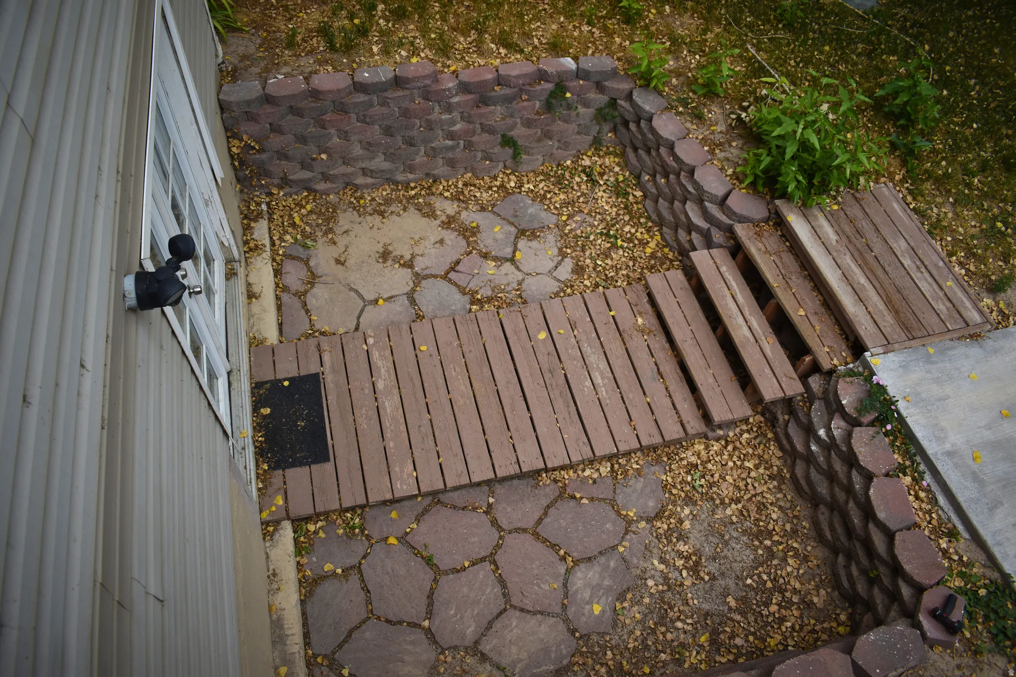 Landscaped outdoor area featuring a patterned paver pathway, wood-plank walking surfaces, and stacked stone retaining walls