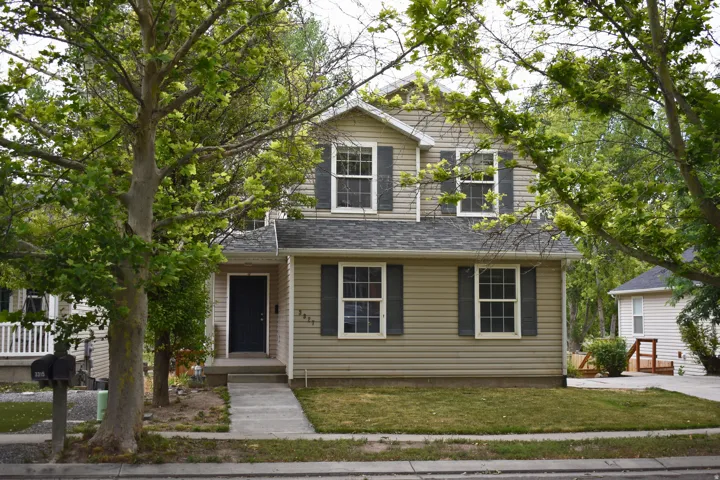 Two-story residence featuring light tan siding, dark shutters, a dark front door, and a gray shingle roof