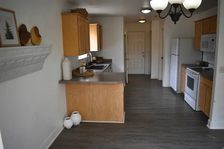 Kitchen area featuring wood-finish flooring, light wood cabinetry, white appliances, dark countertops, and a peninsula with seating space