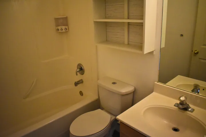 Bathroom featuring a tub/shower combination, white toilet, vanity with integrated sink, and wall-mounted shelving