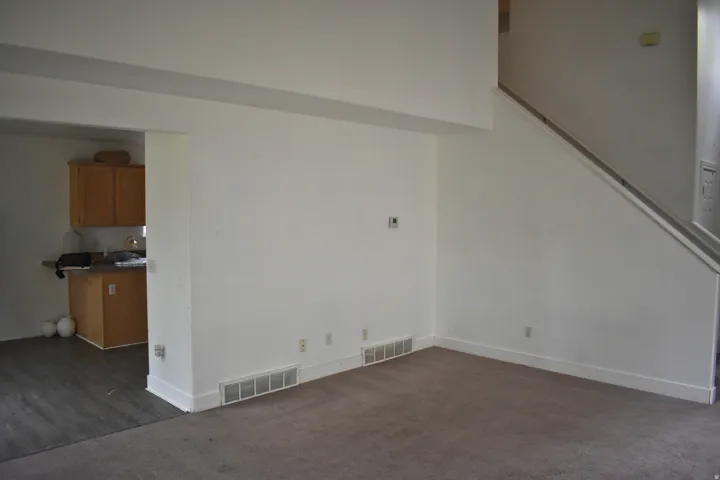 Carpeted living area with light-colored walls, a staircase with a handrail, and a pass-through opening to a kitchen space featuring wood-finish cabinetry, a countertop, and wood-finish flooring