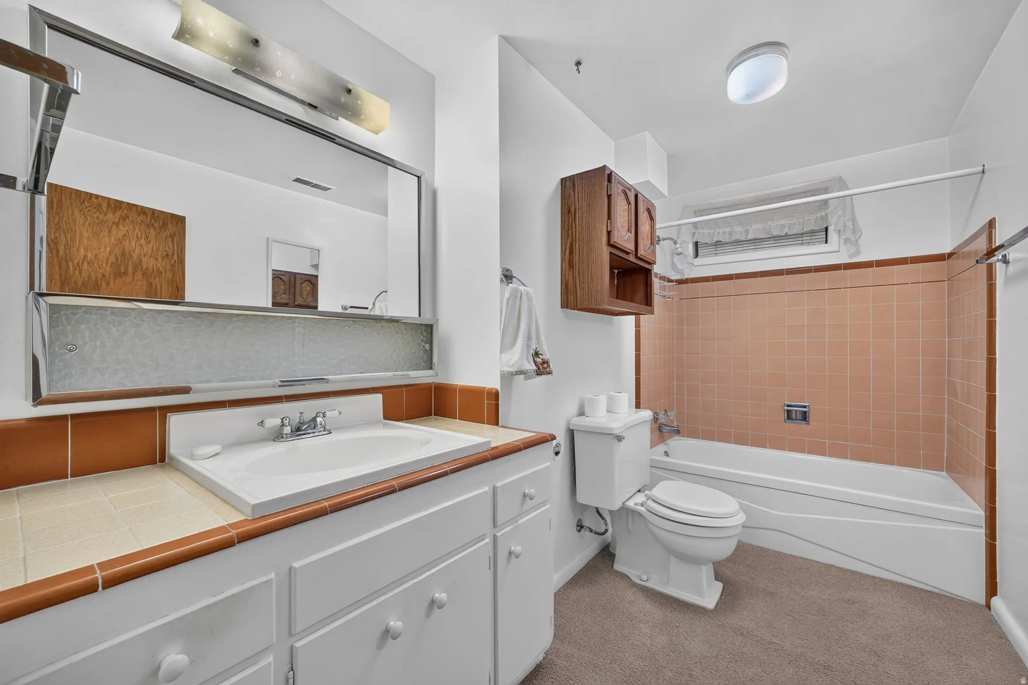 Bathroom featuring a white vanity with integrated sink, a rectangular mirror with integrated lighting, a built-in bathtub with shower, peach-toned tile wainscoting, and a wall-mounted wood cabinet