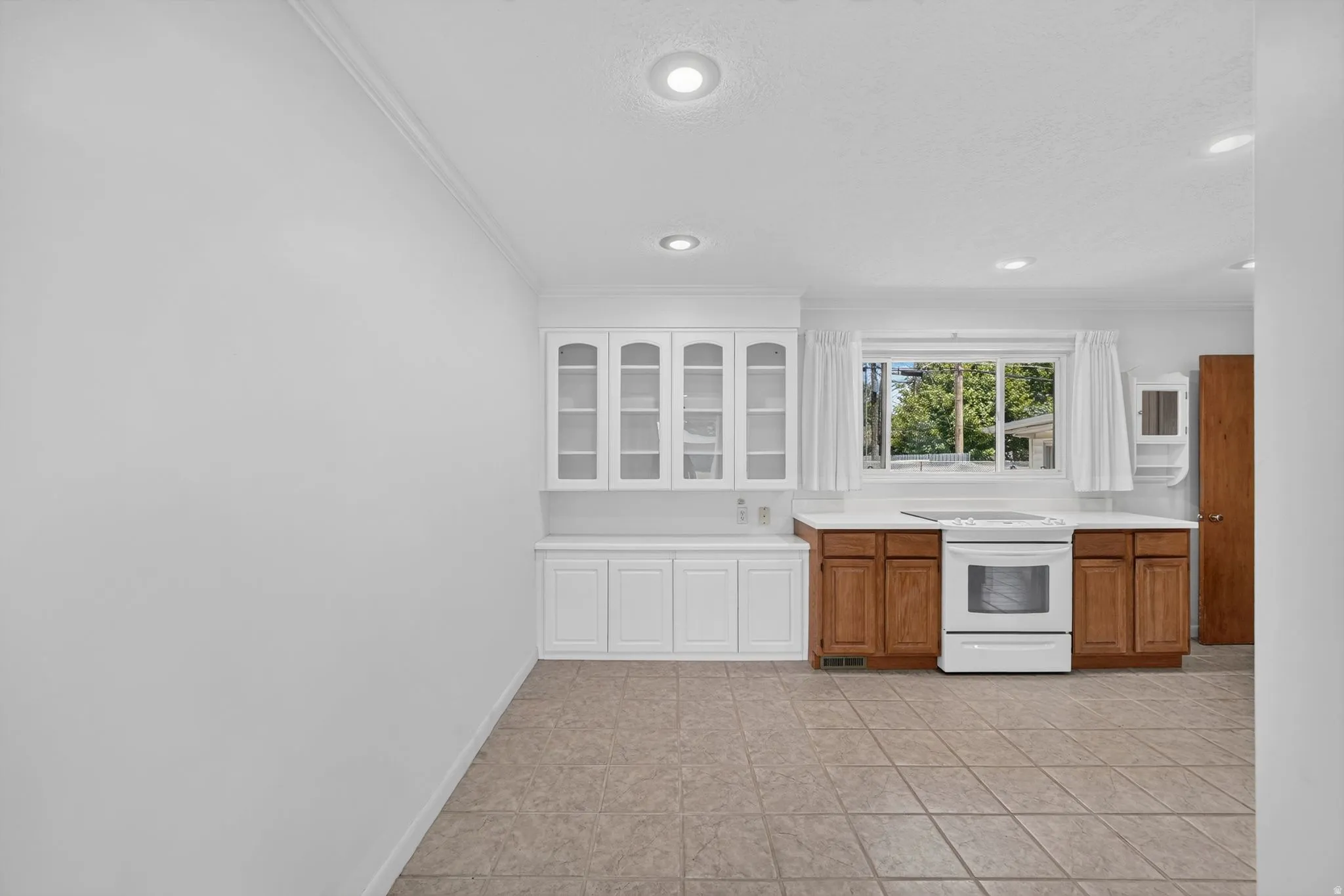 Kitchen area featuring white upper cabinetry with glass inserts, wood-finish lower cabinetry, a white electric range, and a window with exterior views